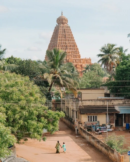 Mother and child walk on dirt road with ancient Indian temple in background, lush trees, rural setting, daytime, clear sky.