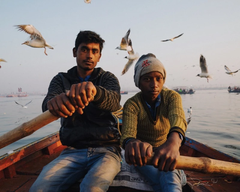 Two people row a wooden boat on a river with many seagulls flying in the sky, captured in a medium shot on a clear day.