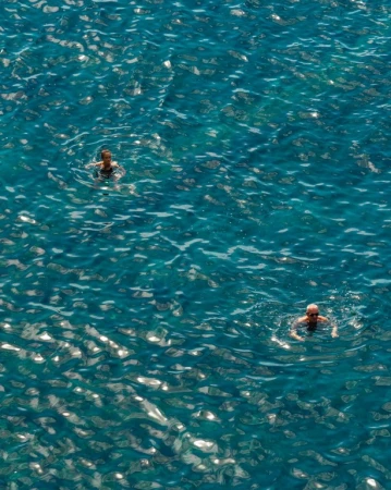 High angle aerial shot of two people swimming in deep blue ocean water with sun glints.