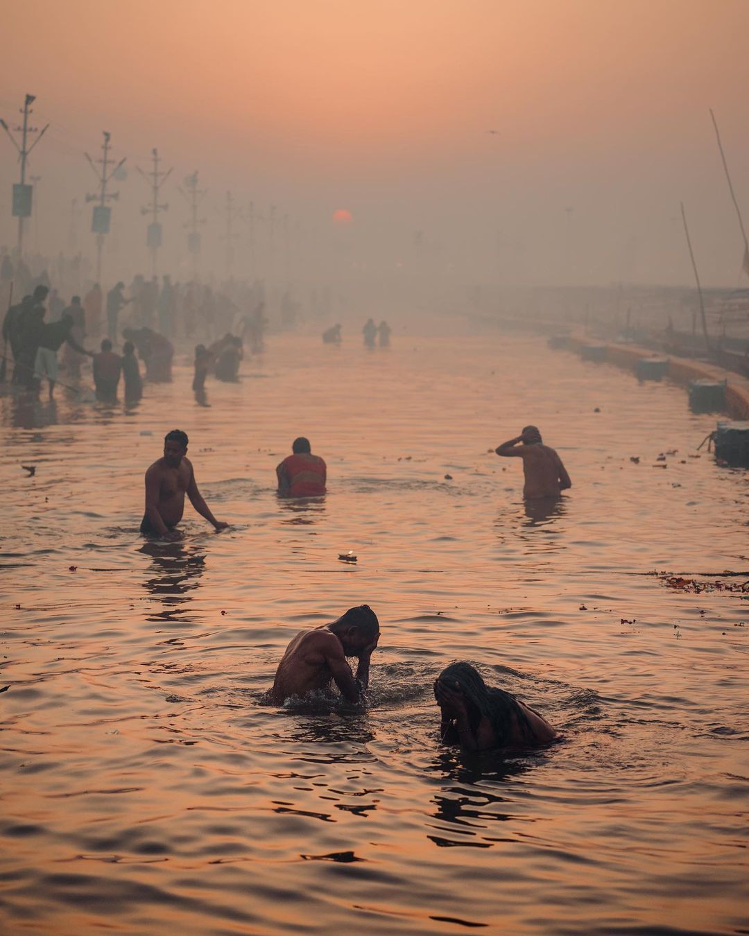 Several partially submerged adult males in a river at sunrise, with a large gathering of people visible in the hazy background.