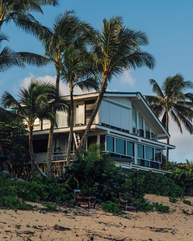 Beach house framed by palm trees under blue sky. Two-story white beach house with rows of windows and lush tropical vegetation.