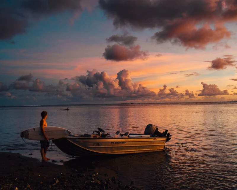 Man with surfboard near motorboat at sunset on beach. Clouds with hues of pink and orange fill the sky. Reflections shimmer on ocean water.