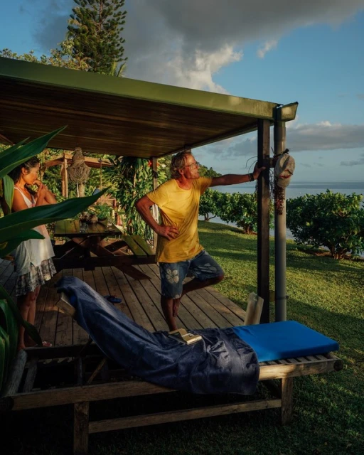 Two adults on a wooden porch, one standing near a post and looking out at the ocean view, the other standing near a table.