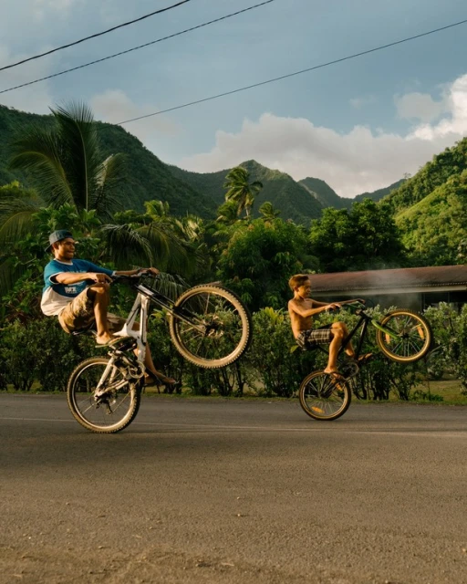 Two people, one adult and one child, perform bicycle stunts on an asphalt road with a lush green mountainous backdrop under a cloudy sky.