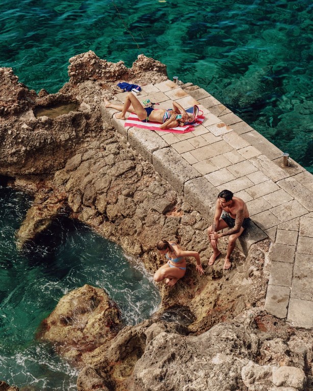 A group of people are enjoying a sunny day by the rocky shore, with one woman tanning on a towel and two others near the clear, turquoise water.
