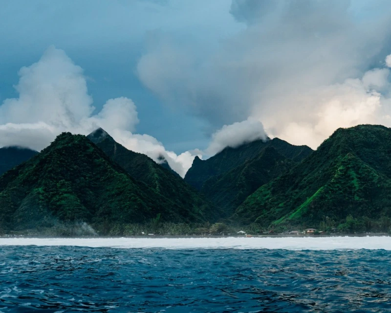 Scenic vista featuring verdant mountain peaks shrouded in clouds, meeting the deep blue ocean under a bright, overcast sky.