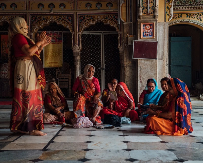 A group of Indian women are sitting on a marble floor inside a building, some reading books, dressed in traditional clothing with headscarves.