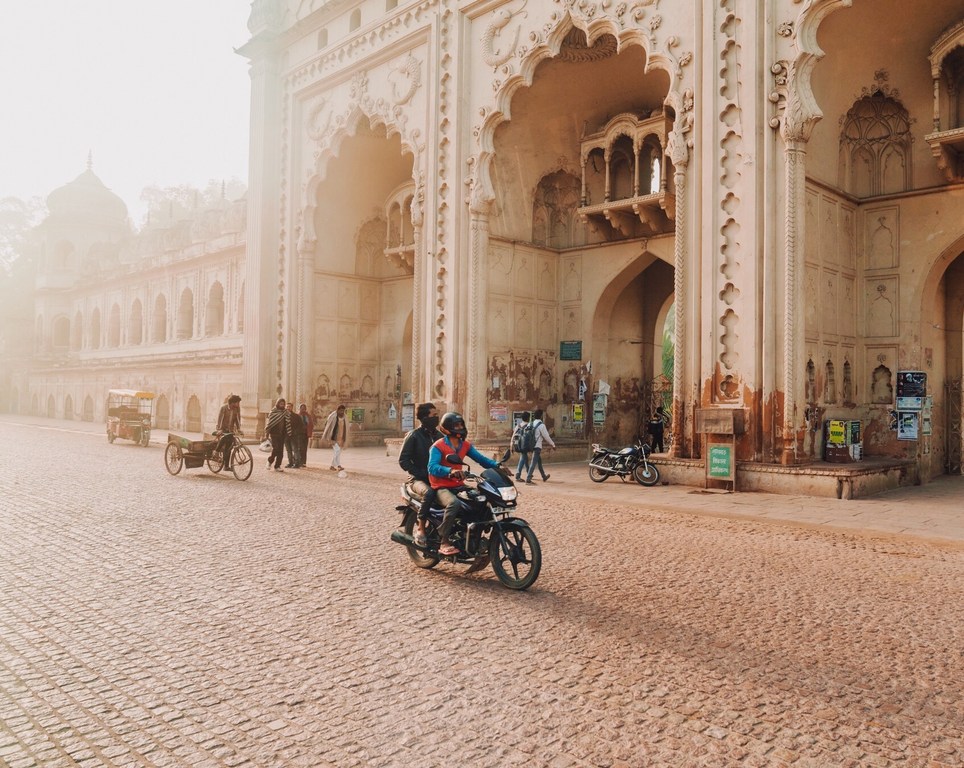 People on a motorcycle ride past the architectural gate in Lucknow, India on a cobblestone street during a sunny day.