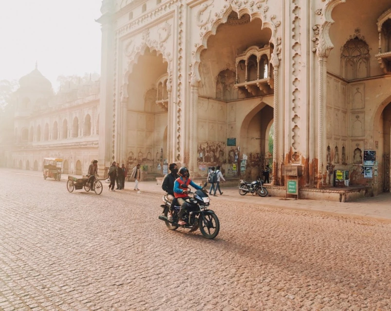 People on a motorcycle ride past the architectural gate in Lucknow, India on a cobblestone street during a sunny day.
