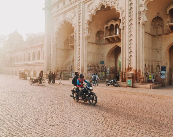 People on a motorcycle ride past the architectural gate in Lucknow, India on a cobblestone street during a sunny day.