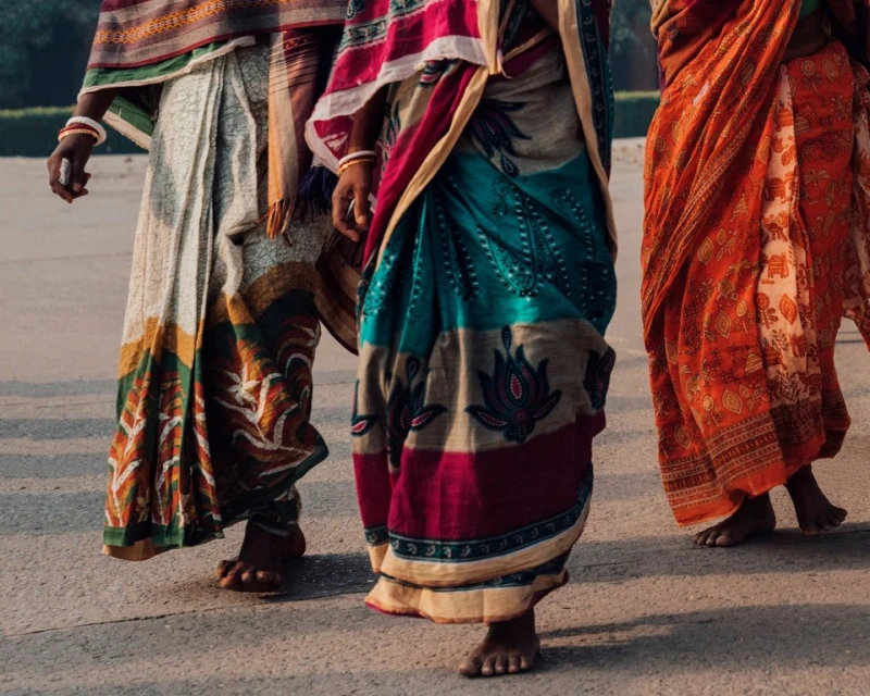 Three women walk in colorful saris, showcasing traditional Indian fashion with intricate patterns and vibrant colors on a sunny day.