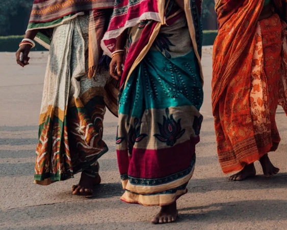 Three women walk in colorful saris, showcasing traditional Indian fashion with intricate patterns and vibrant colors on a sunny day.