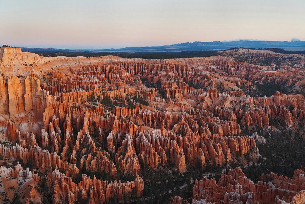 Sweeping vista of Bryce Canyon's orange hoodoos stretching to distant mountains under soft sky at sunrise; landscape photography.