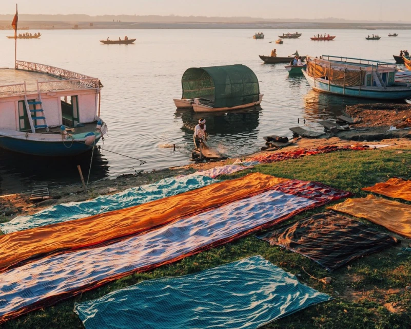 Indian fisherman stands in water by boats with drying colorful textiles, on a riverbank at sunrise with clear sky in Varanasi, India.