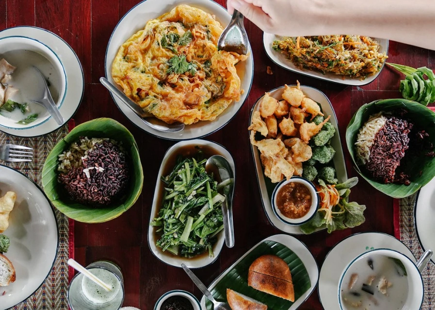 Overhead shot of a variety of Thai dishes, including omelets, rice, vegetables, and soup, served on a dark wooden table.