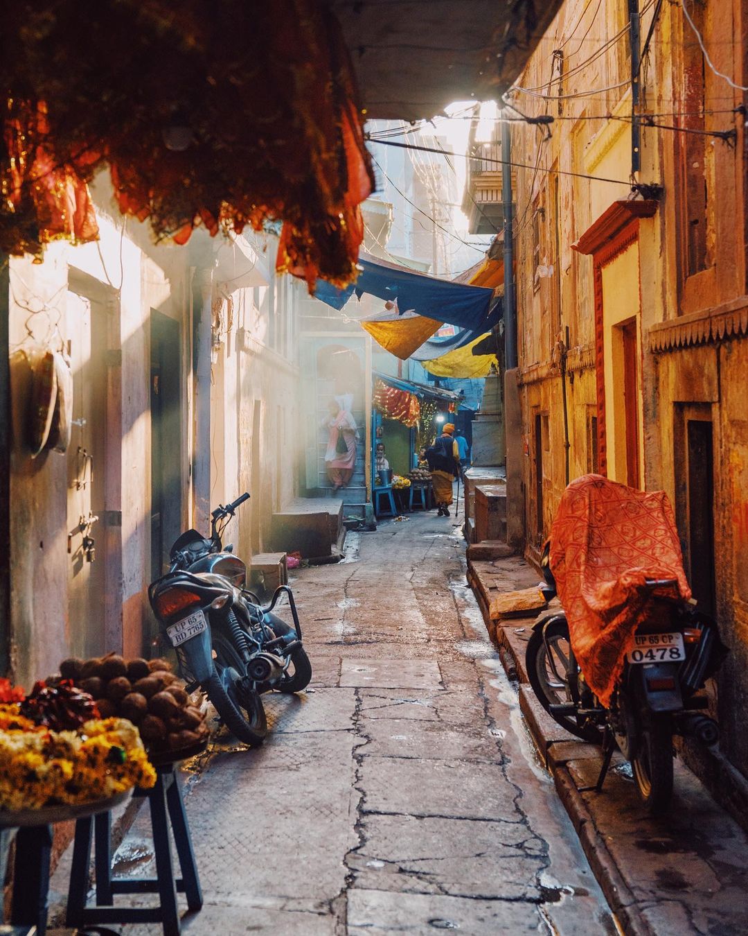 Urban scene of a narrow alleyway in India with motorbikes, draped textiles, a distant person walking away, and soft, warm, natural lighting.