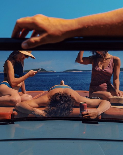 Four people relax on a boat deck, with a close-up view of a hand holding the top of the windshield, islands, and a blue sea visible.