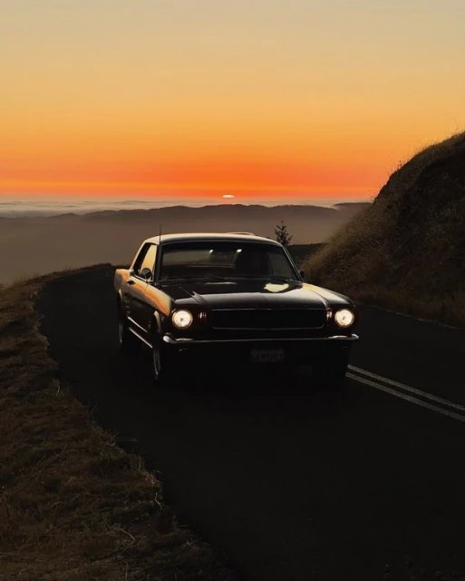Vintage black Ford Mustang car driving along a winding road at sunset with orange sky and mountains in the distance.