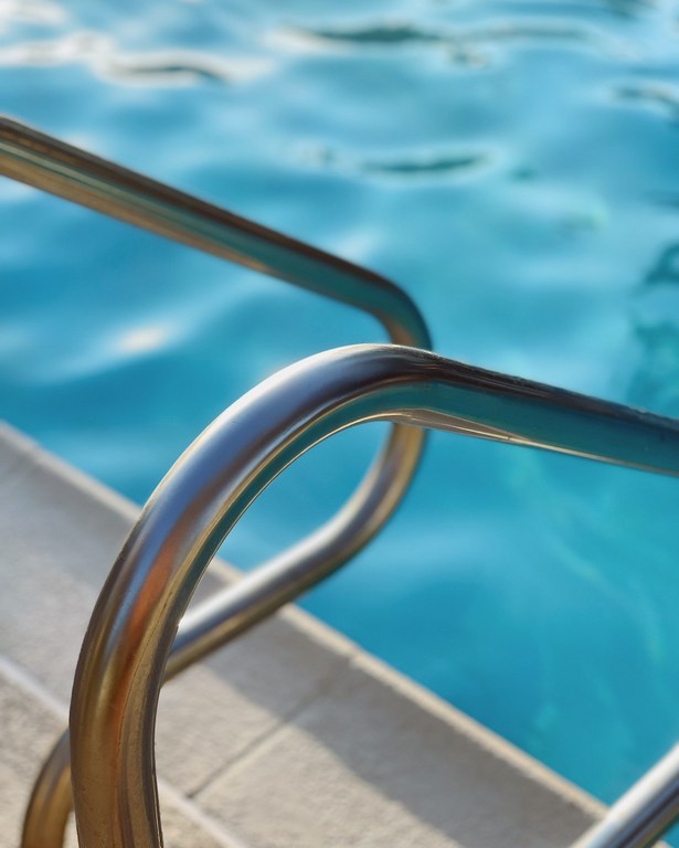 Close-up of stainless steel pool ladder steps going into blue swimming pool water, shot with shallow depth of field on a sunny day.