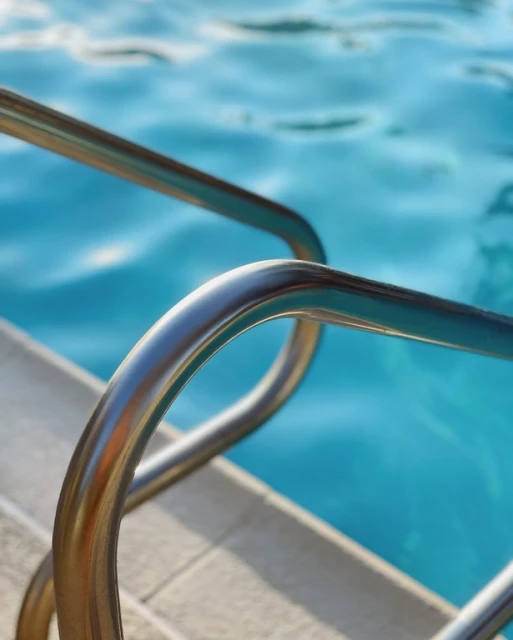Close-up of stainless steel pool ladder steps going into blue swimming pool water, shot with shallow depth of field on a sunny day.