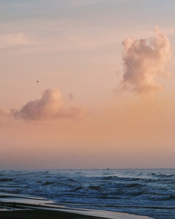 Calm ocean waves under a pastel sunset sky with soft clouds, gentle shoreline, and distant boat on the horizon create a serene seascape.