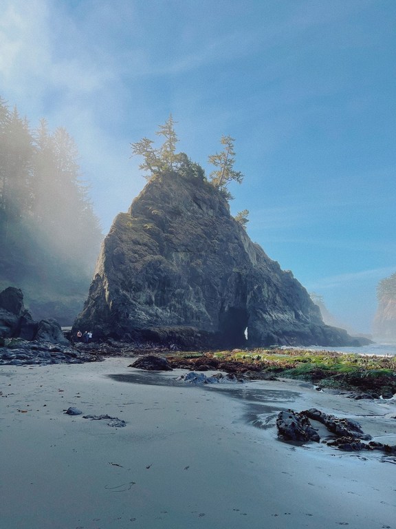 Scenic view of a dramatic rocky sea stack with trees on top at a beach on a misty day with blue sky and grey sand.
