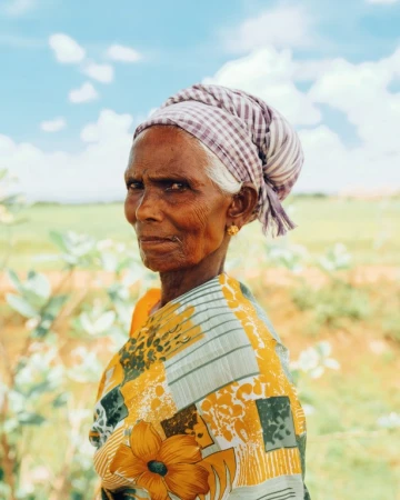 Elderly Indian woman in patterned headscarf and dress stands outdoors against a blue sky with light clouds and a green, grassy background.