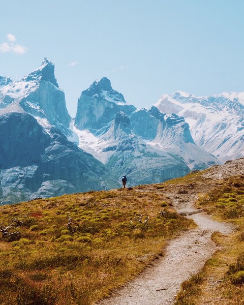 Hiker in a blue jacket walks a dirt path with snow-capped mountains in the background under a clear blue sky on a sunny day.