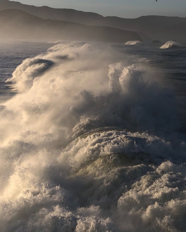 Waves crashing on the ocean with mountains in the background. Spray and mist fill the air, creating a dynamic coastal landscape.