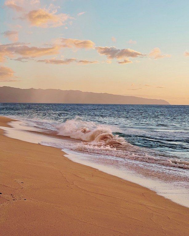 Ocean waves washing onto a tan sandy beach, with a mountain range visible in the background under a pastel sunset sky.