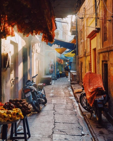 Narrow sunny street scene with parked motorcycles, draped fabrics above, and a person walking in the distance, captured by Dan Tom