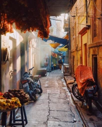 Narrow sunny street scene with parked motorcycles, draped fabrics above, and a person walking in the distance, captured by Dan Tom