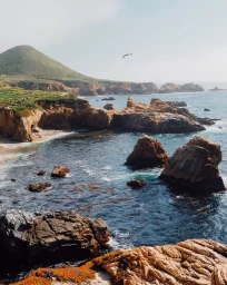 Coastal landscape photography by Dan Tom featuring rocky cliffs meeting the ocean with a green hill and flying birds above
