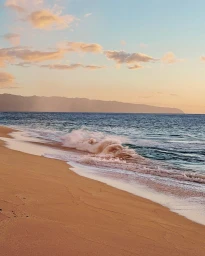 Coastal landscape with gentle waves breaking on a sandy beach under a soft sunset sky with distant mountains.