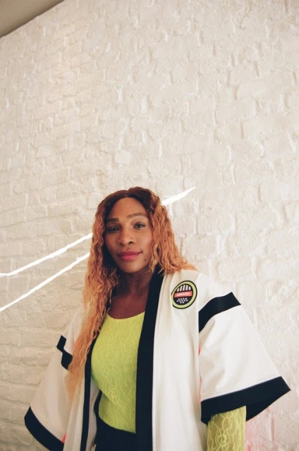 Serena Williams poses in a white jacket with black trim against a white brick wall, wearing a yellow lace top and black bottoms.