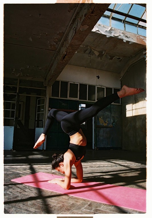 Woman in black sportswear doing a handstand on a pink yoga mat in an abandoned industrial interior under sunlight.