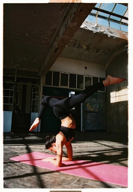 Woman in black sportswear doing a handstand on a pink yoga mat in an abandoned industrial interior under sunlight.