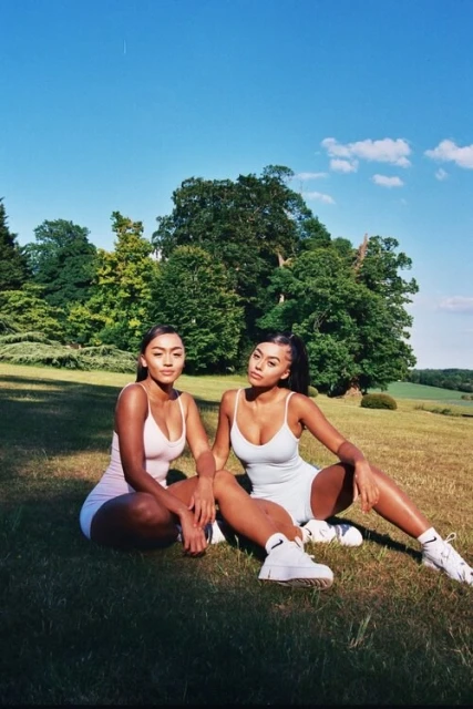 Two women pose in athletic wear on a lawn, smiling against a backdrop of green trees and a bright blue sky on a sunny day.