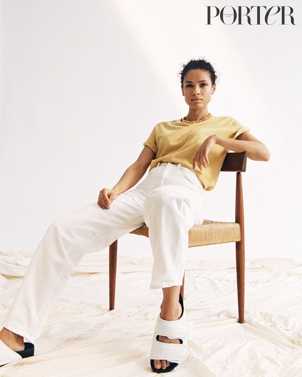 A young adult woman sits on a chair in a photo studio, wearing a yellow t-shirt and white pants, with studio lighting.