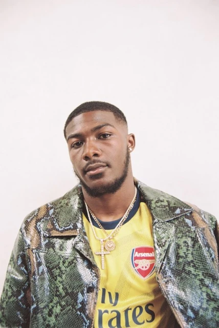 Portrait of a young Black man wearing an Arsenal soccer jersey and snake-print jacket against a white background in eye-level shot.