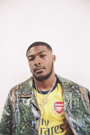 Portrait of a young Black man wearing an Arsenal soccer jersey and snake-print jacket against a white background in eye-level shot.