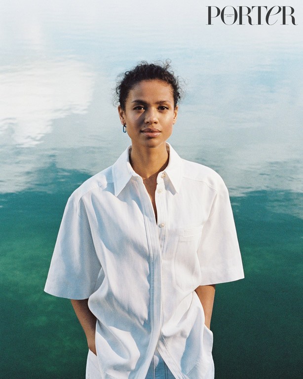 Woman in white shirt poses in front of water for a portrait in front of light teal water reflecting the sky, with 'PORTER' logo visible.