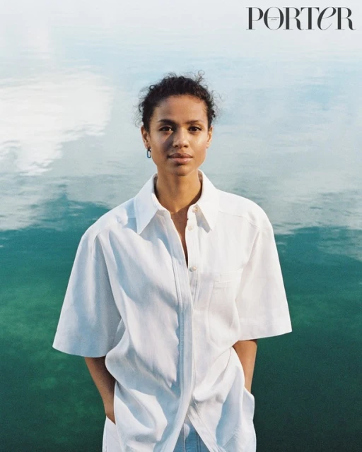 Woman in white shirt poses in front of water for a portrait in front of light teal water reflecting the sky, with 'PORTER' logo visible.