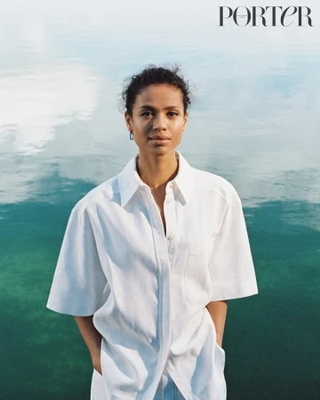 Woman in white shirt poses in front of water for a portrait in front of light teal water reflecting the sky, with 'PORTER' logo visible.