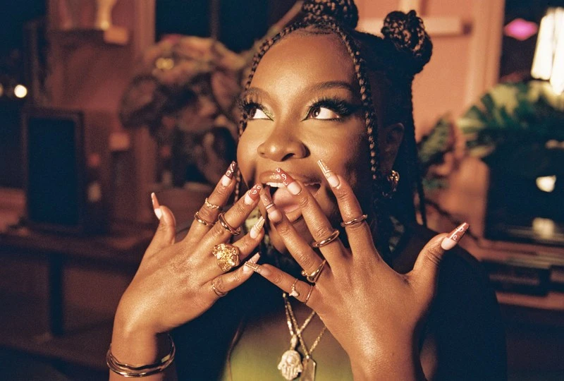 Woman with elaborate nails and jewelry posing indoors, with decorative plants and interior lighting in the background creating warm tones.