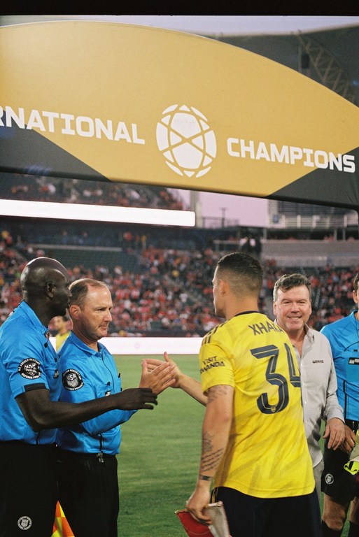 Soccer player in yellow jersey shakes hands with referee on field during International Champions Cup, crowd and sign in background.