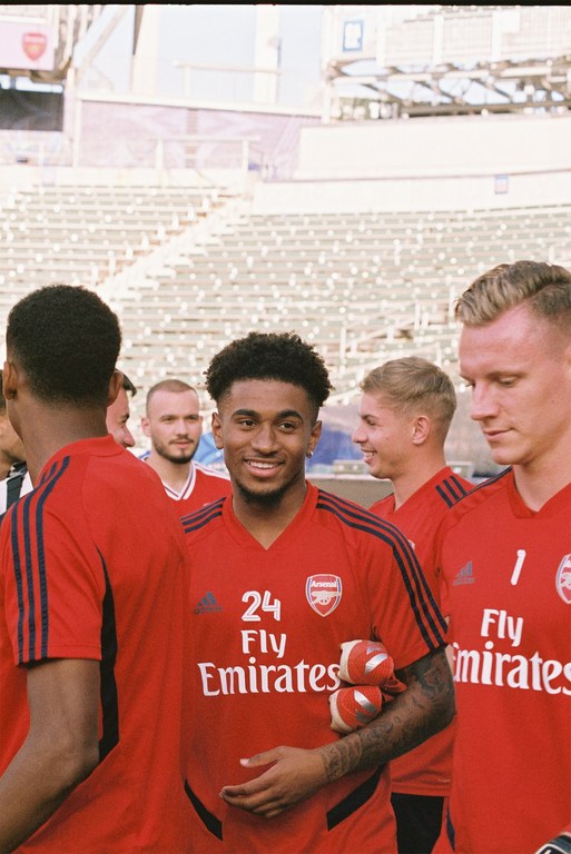 Group of soccer players in red jerseys with 'Arsenal Fly Emirates' on the front, smiling on a stadium field, medium outdoor shot.