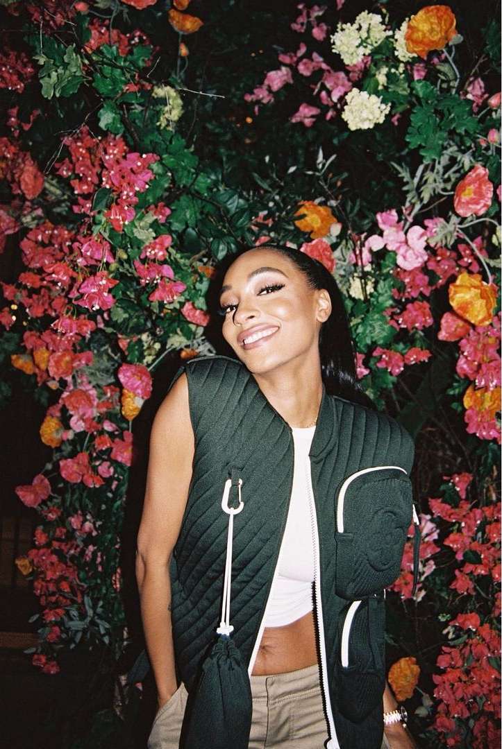 Smiling woman poses in front of a floral background wearing a puffy vest, top, and pants. Close-up shot in outdoor setting.