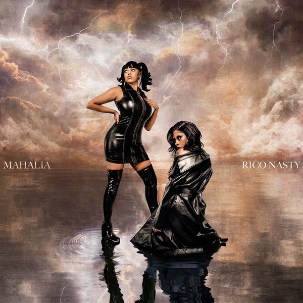 Two women pose in shiny black outfits against a dramatic sky backdrop, reflected on a water-like surface, evoking a bold, artistic aesthetic.