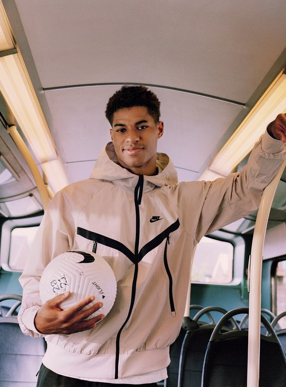 Young man in hooded jacket poses inside a bus, holding a soccer ball with the Nike logo; indoor setting with natural light.